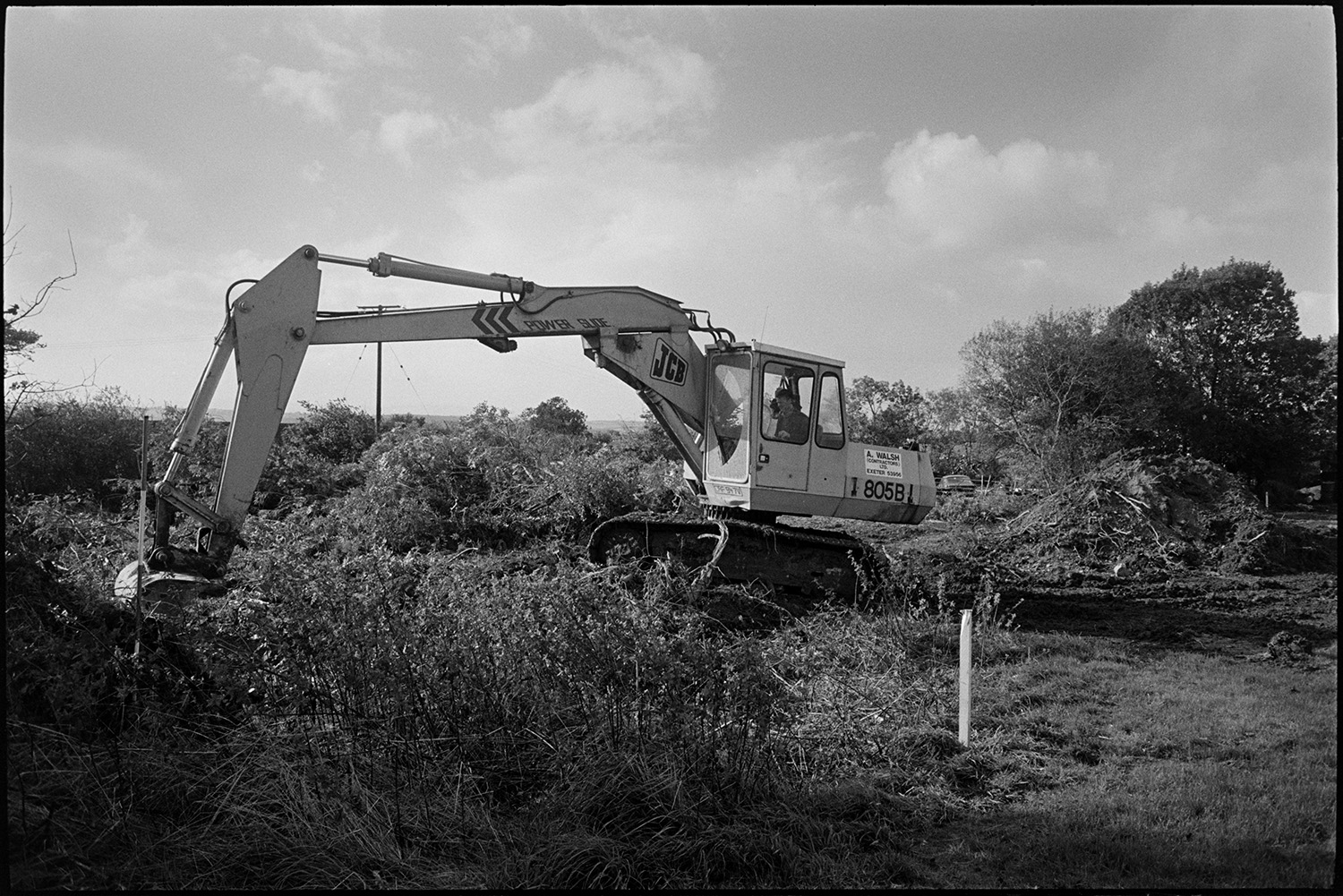 Builders starting work on site of new school, excavator and men drilling first hole on site.
[Contractors making a start on the site of the new primary school in Chulmleigh. A mechanical JCB digger is removing a hedge for the groundworks.]