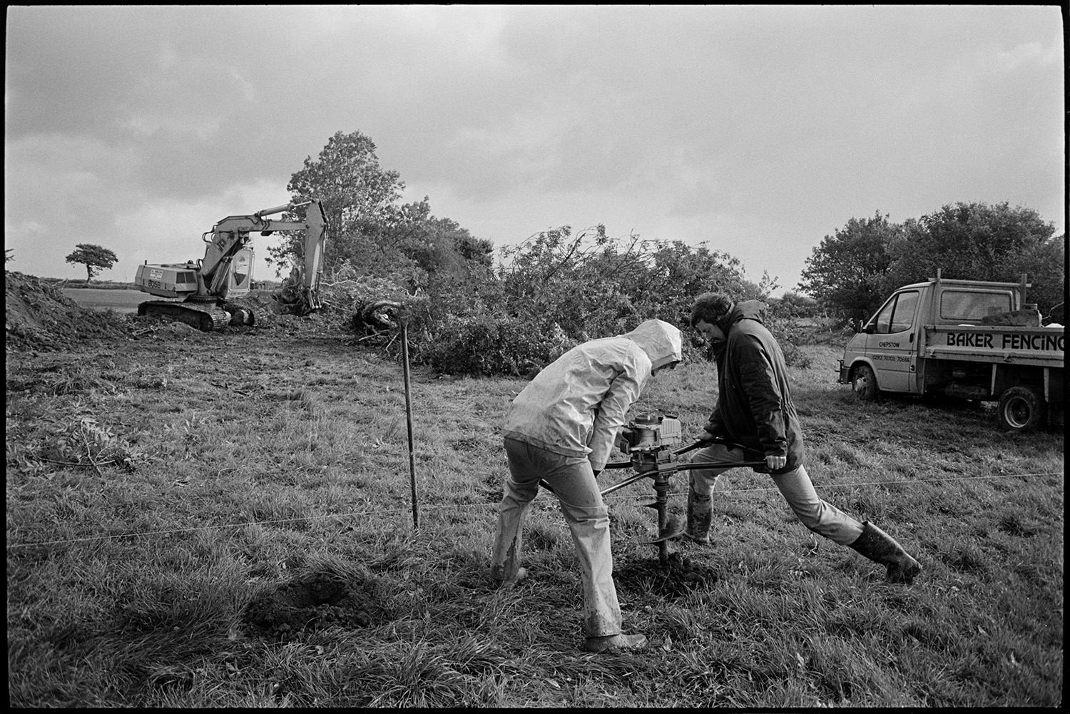 Excavator clearing ground for new school buildings, men drilling.
[Two men using a hole borer on the site of the new primary school at Chulmleigh. A mechanical digger is clearing a hedge in the background, alongside a vehicle belonging to Baker Fencing.]