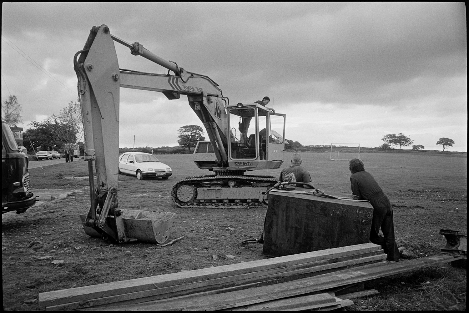 Excavator clearing ground for new school buildings, men drilling.
[Two men standing by a metal tank and planks of wood, near a JCB digger on the site of the new primary school at Chulmleigh. There is another man standing on the excavator. They are looking towards a sports field, cars and a bus in the background.]
