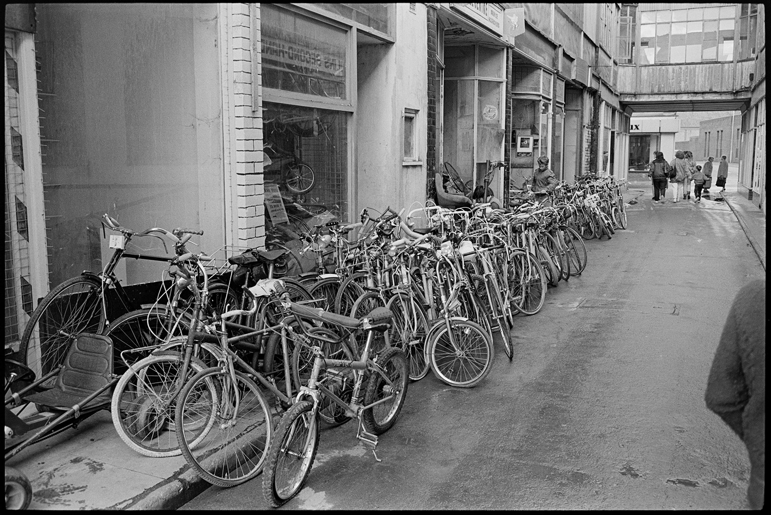 Rows of bicycles stacked outside shop fronts in a shopping arcade off Bear Street, Barnstaple. There are pedestrians at the end of the arcade.