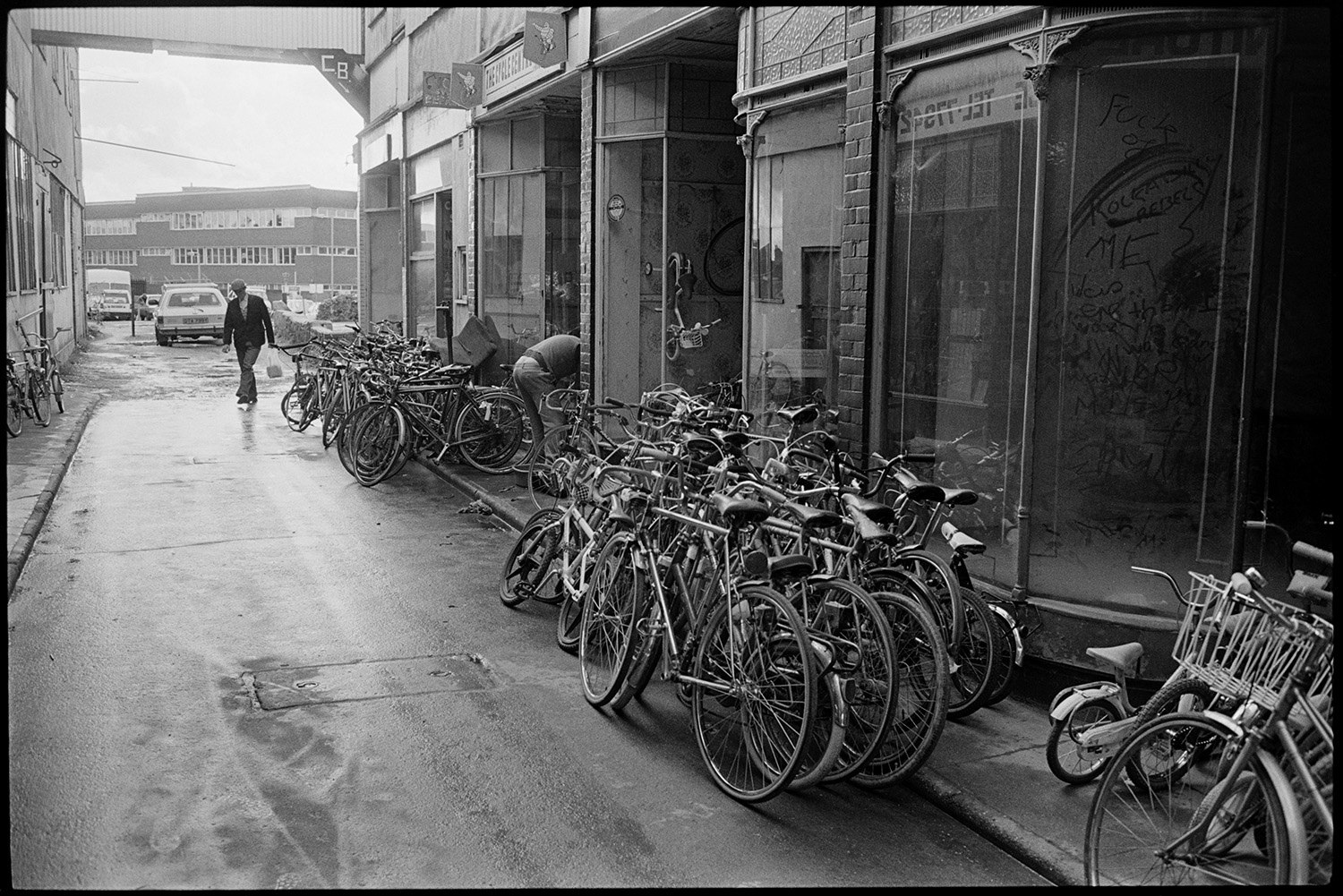 Rows of bicycles stacked outside shop fronts in a shopping arcade off Bear Street, Barnstaple. Some of the shops are empty. A man is walking into the arcade. Parked cars and an office block are visible in the background.