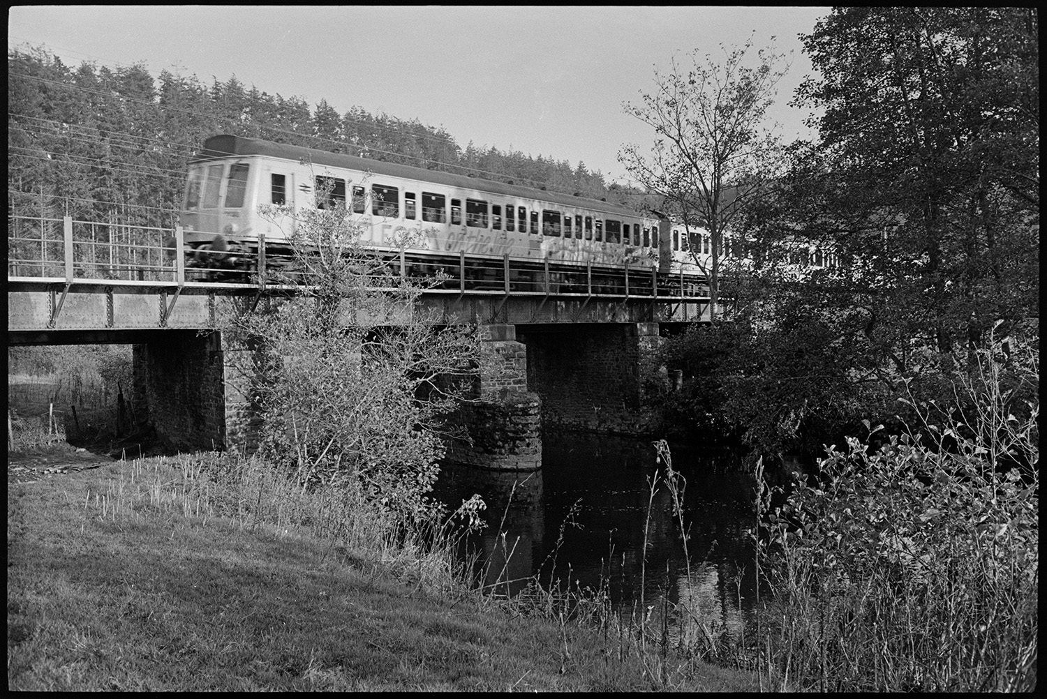 Railway bridge over river, men repairing bridge, walkway.
[A diesel train crossing a railway bridge over the River Taw near Eggesford. The bridge is supported by stone pillars.]