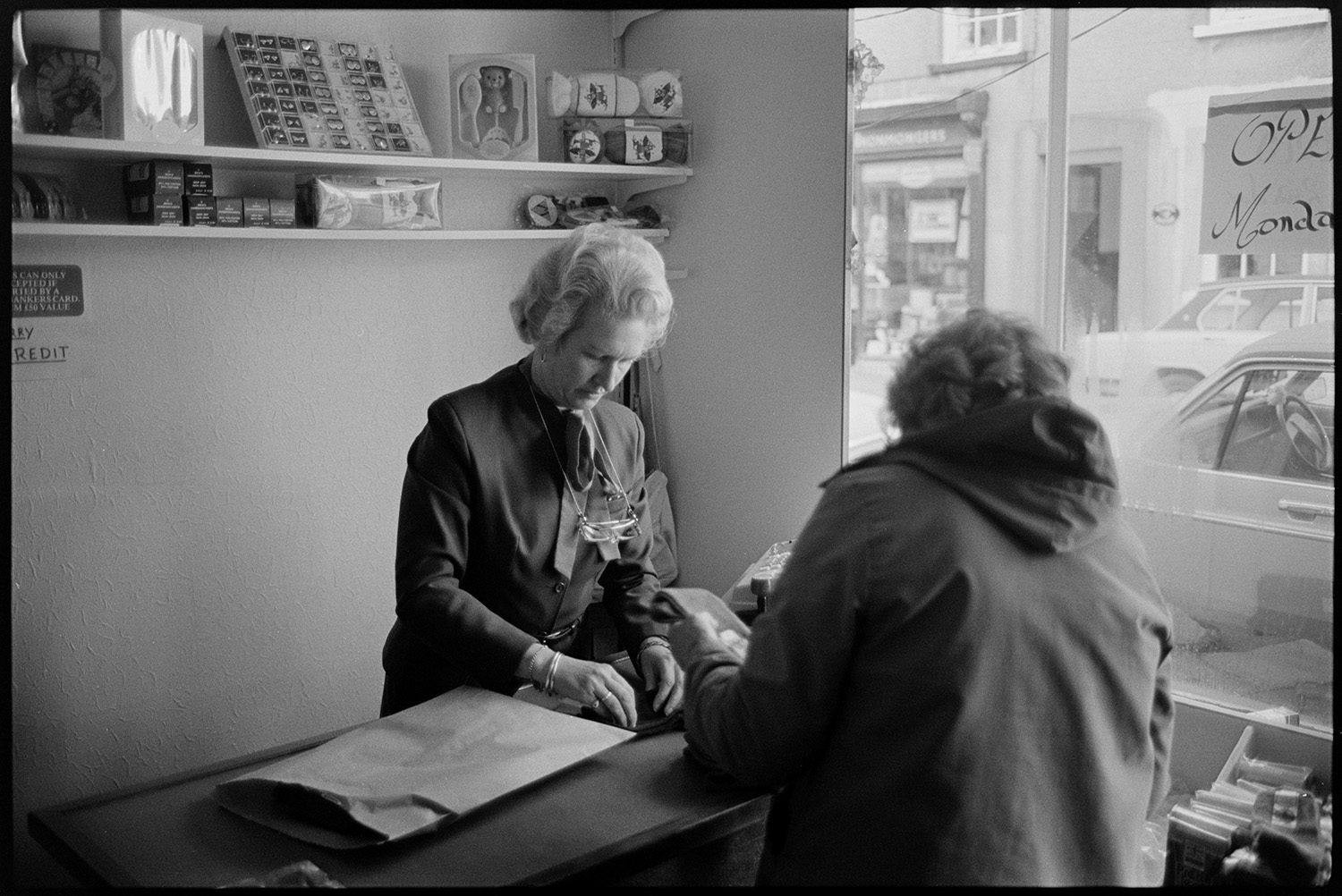 Clothes shop, woman proprietor serving customer on first day of business.
[A woman proprietor serving a customer on her first day of business in Birkett's clothes shop in Chulmleigh. Gifts, including jewellery, are displayed on shelves behind the counter. Through the shop window can be seen parked cars on Fore Street.]
