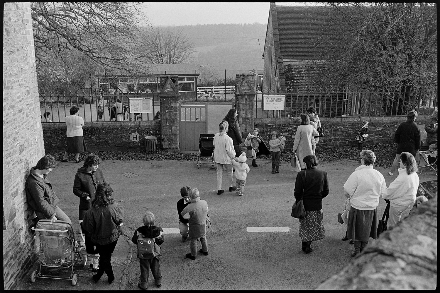 Women, mothers waiting for their children outside school.
[Mothers waiting for their children at the gates of the old Chulmleigh Primary School. Women and children are standing in the street, with the school playground and school buildings in the background.]