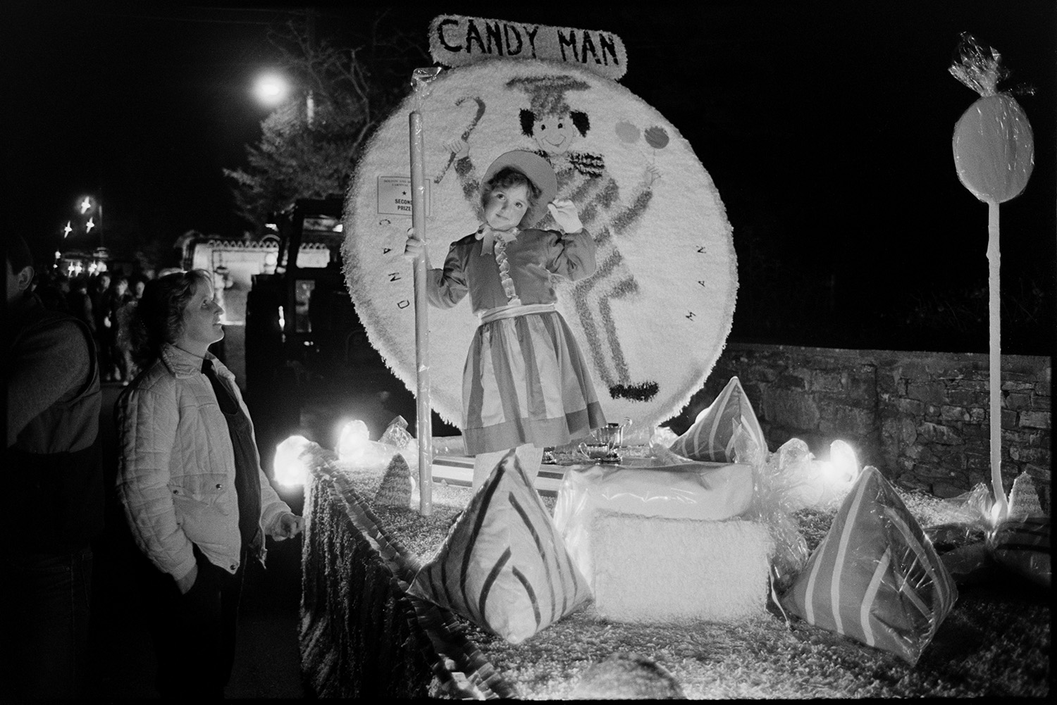 Carnival floats at night, Queen and attendants, fancy dress.
[A carnival float during the evening procession at Dolton Carnival. A young girl is standing in fancy dress on a float called Candy Man. A woman stands beside the decorated float, with lights and spectators in the background.]