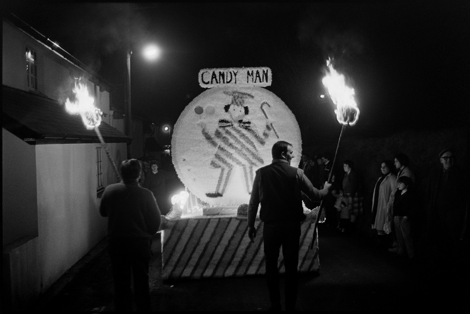 Carnival floats at night, queen and attendants, fancy dress with burning torches.
[A carnival float during the evening procession at Dolton Carnival. Two men are walking with burning torches behind a carnival float called 'Candy Man'. Spectators are watching the procession.]