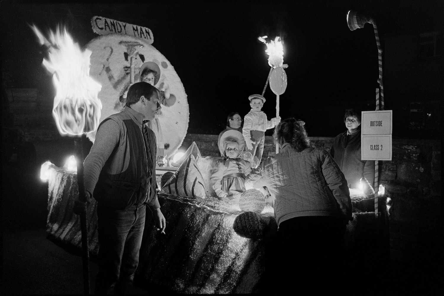 Carnival floats at night, queen and attendants, fancy dress with burning torches.
[A carnival float during the evening procession at Dolton Carnival. Two girls and boy are in fancy dress and on a float called 'Candy Man'. Men and women are standing around the float, with lights and burning torches.]