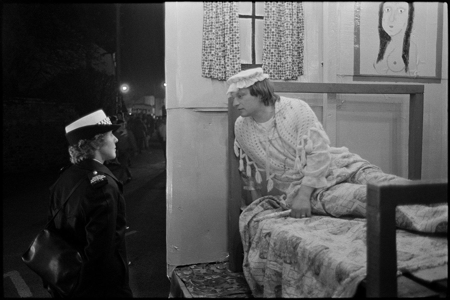 Carnival floats at night, queen and attendants, fancy dress.
[A carnival float during the evening procession at Dolton Carnival. A young man in fancy dress is lying on a bed and talking to a policewoman standing by the carnival float.]