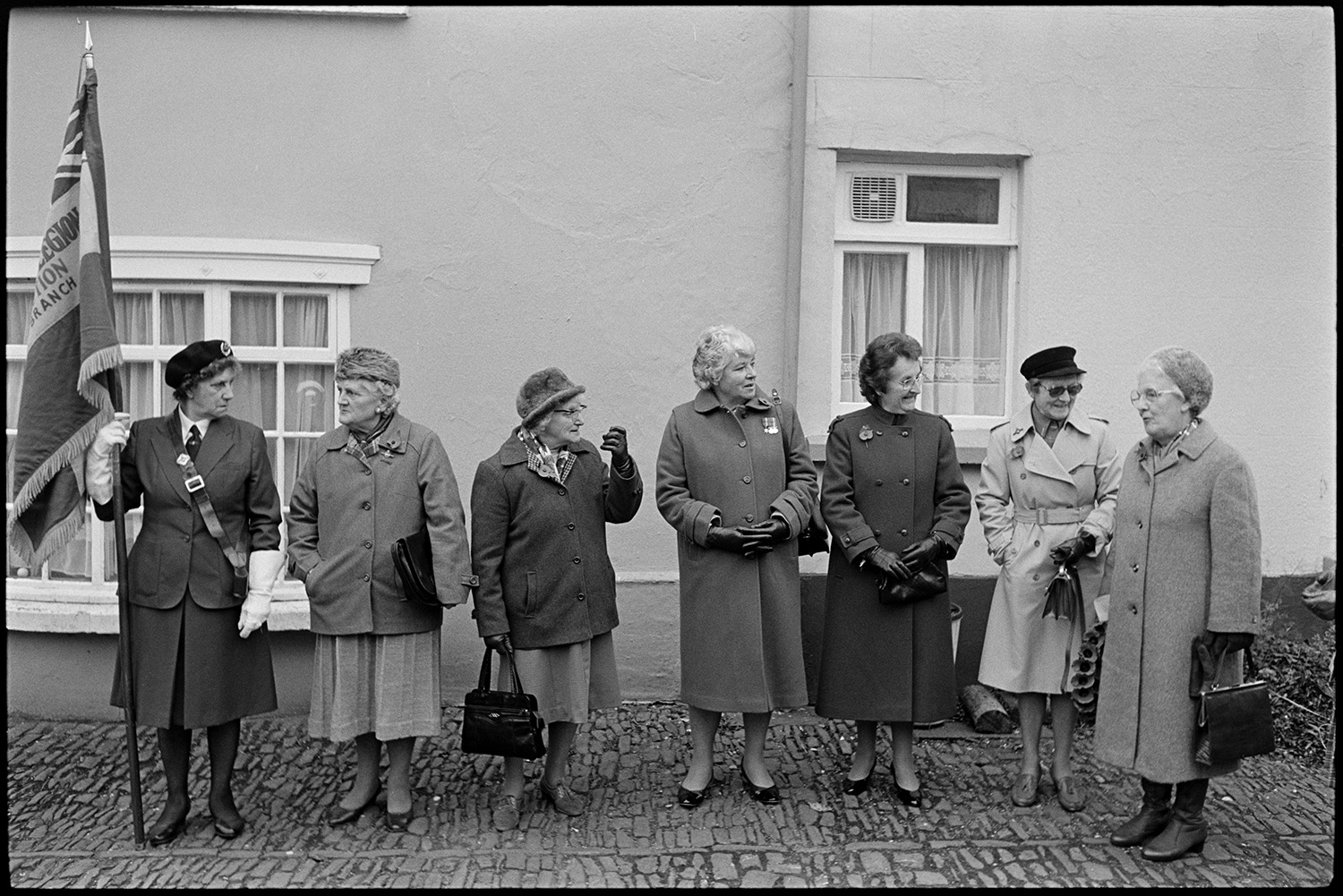 Men and women assembling for Remembrance Sunday Parade, some with bowler hats and medals.
[Seven woman waiting for the Remembrance Sunday Parade at Chulmleigh. They are standing on a cobbled pavement in front of a house. One is in uniform and holding a Royal British Legion flag, another is wearing medals and the other women are wearing poppies.]