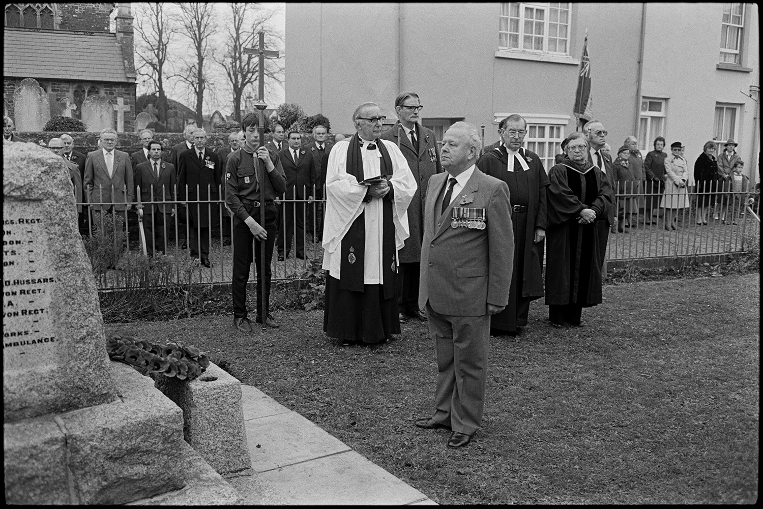 Parade at Memorial, laying wreaths by men, women and scouts. Vicar saying prayers.
[Men and women assembling for the Remembrance Sunday wreath laying ceremony at the war memorial in the square at Chulmleigh. Some men are wearing medals. The Reverend John Richards is taking the service and a man has just laid a wreath. A scout carries a cross, and a woman carries a British Legion flag in the background.]