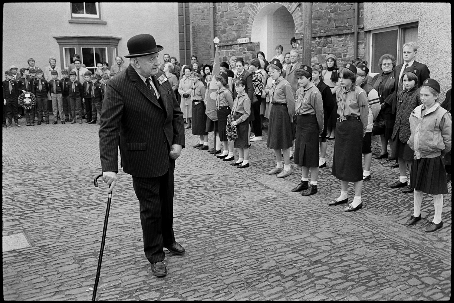 Parade at Memorial, laying wreaths, by men, women and scouts. Vicar saying prayers.
[People assembling for the Remembrance Sunday Parade on the cobbled square in Chulmleigh. One man is inspecting groups of Scouts and Girl Guides. He is wearing a bowler hat, a suit and medals.]