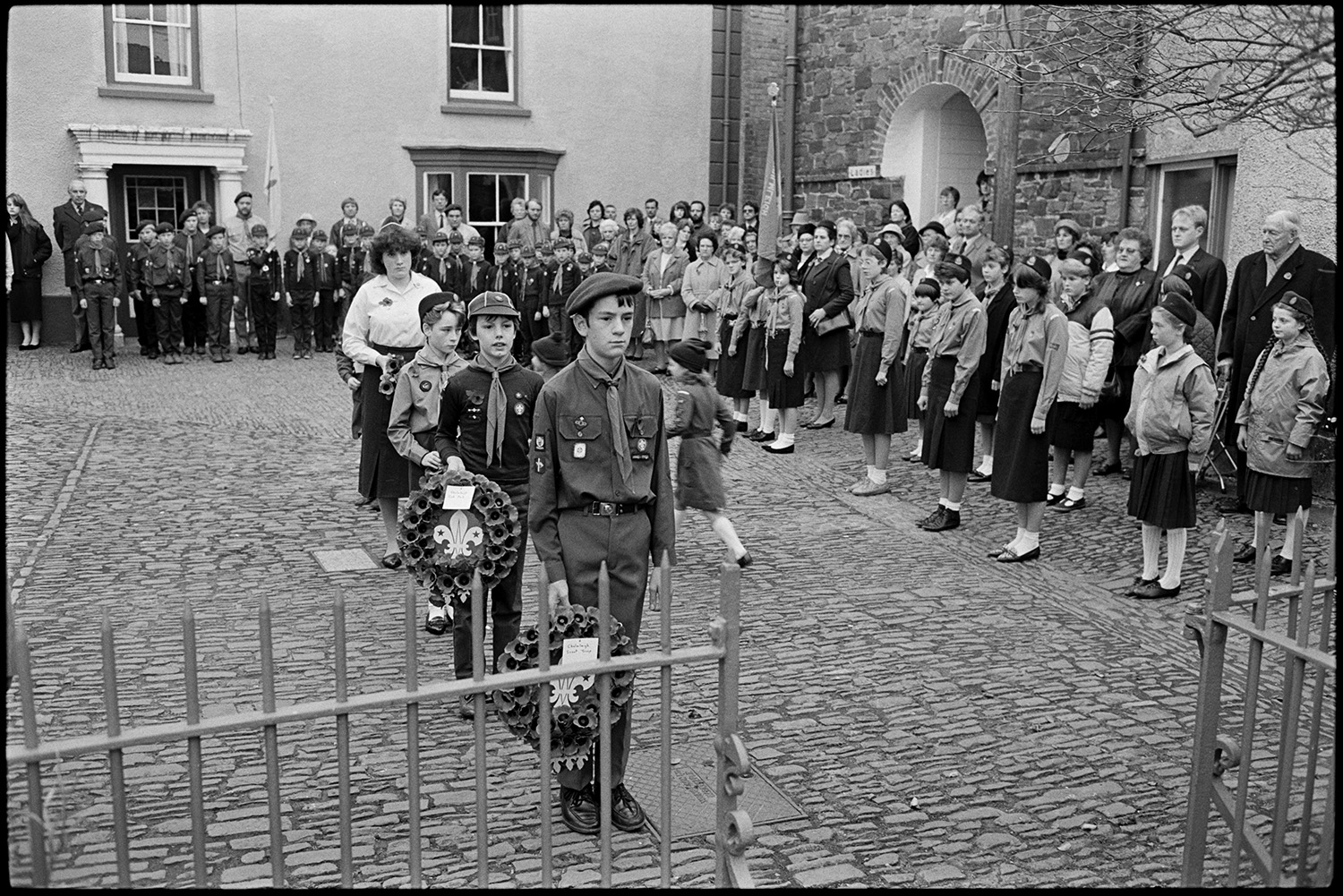 Parade at Memorial, laying wreaths by men, women and scouts. Vicar saying prayers.
[People assembling for the Remembrance Sunday wreath laying ceremony at the war memorial in the cobbled square at Chulmleigh. There are groups of Girl Guides and Scouts. Two Scouts are carrying poppy wreaths to the war memorial.]