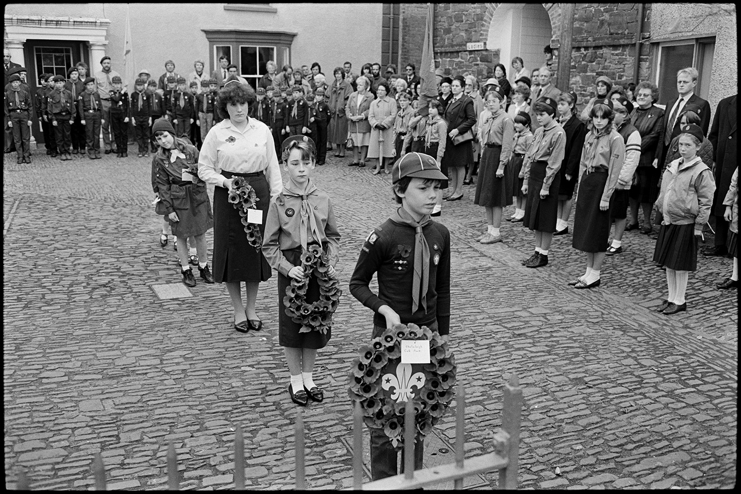 Parade at Memorial, laying wreaths by men, women and scouts. Vicar saying prayers.
[People assembling for the Remembrance Sunday wreath laying ceremony at the war memorial in the cobbled square at Chulmleigh. There are groups of Girl Guides and Scouts. One Scout, a Girl Guide, a Guide leader and a Brownie are carrying wreaths to the memorial.]