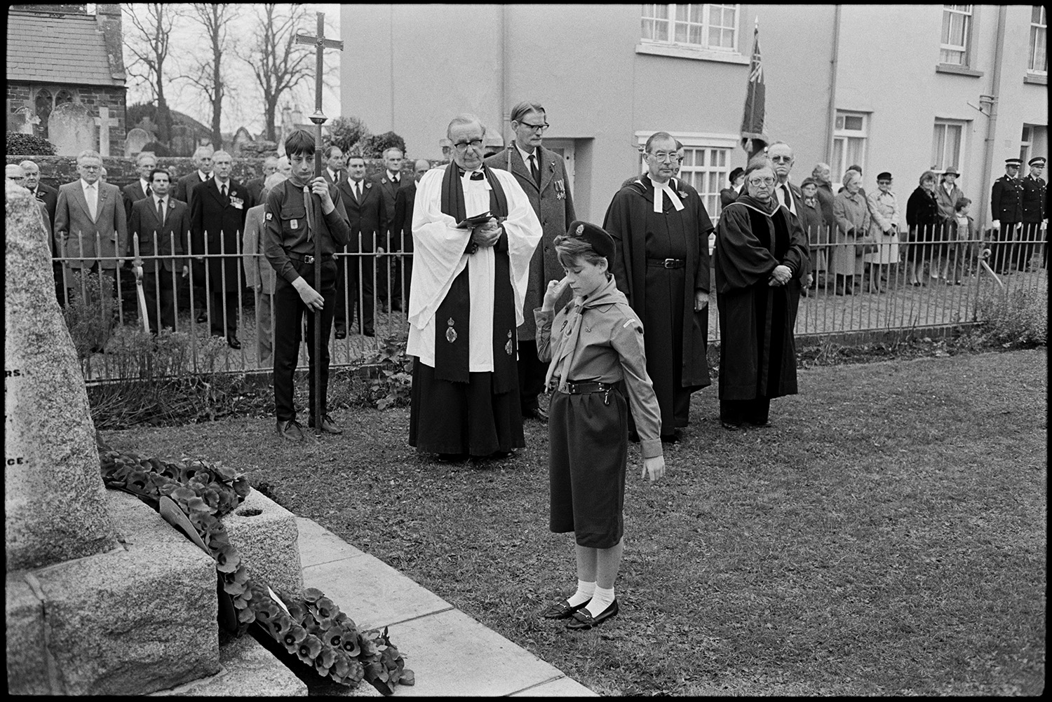 Parade at Memorial, laying wreaths by men, women and scouts. Vicar saying prayers.
[People assembling for the Remembrance Sunday wreath laying ceremony at the war memorial in the square at Chulmleigh. The Reverend John Richards is taking the service. A Girl Guide salutes after laying a wreath on the memorial step. IN the background a scout carries a cross, and a woman carries a Royal British Legion flag. ]