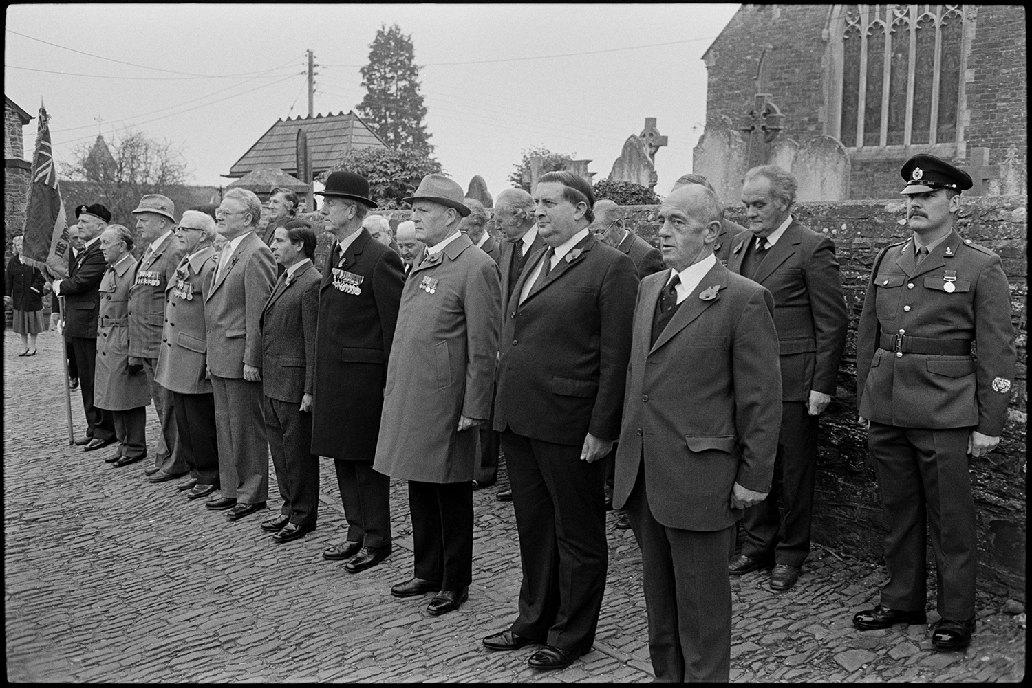 Parade at Memorial, laying wreaths by men, women and scouts, Vicar saying prayers.
[Men standing on a cobbled path outside Chulmleigh Church for the two minute silence on Remembrance Sunday or Armistice Day. They are wearing poppies and some of them are wearing medals and uniforms.]