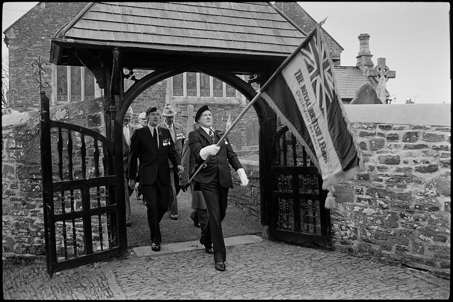 Parade at War Memorial, two minute silence, march to church, chatting afterwards, scouts.
[A group of men coming through the Lych Gate at Chulmleigh Church during Remembrance Sunday or Armistice Day to lead a procession to the war memorial. One is carrying the Royal British Legion flag, they are wearing poppies and some of them are wearing medals.]