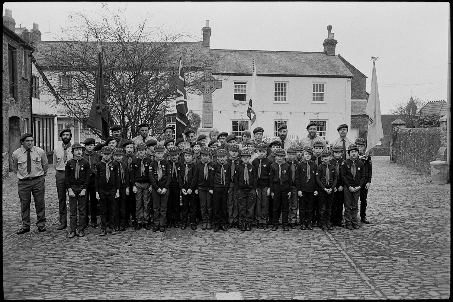 Parade at War Memorial, two minute silence, march to church, chatting afterwards, scouts.
[A group of Scouts with their leaders, standing in the cobbled town square in Chulmleigh, during Remembrance Sunday or Armistice Day. They are holding up four flags, and standing in front of the War Memorial, with town houses in the background.]
