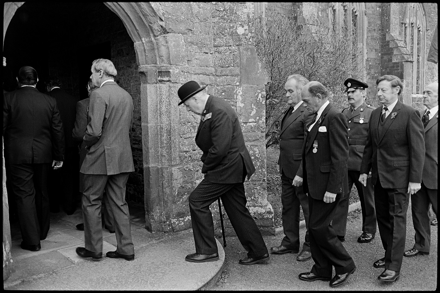 Parade at War Memorial, two minute silence, march to church, chatting afterwards, scouts.
[Men entering Chulmleigh Church during Remembrance Sunday or Armistice Day. They are wearing poppies and some of them are wearing medals. One man is wearing a military uniform.]