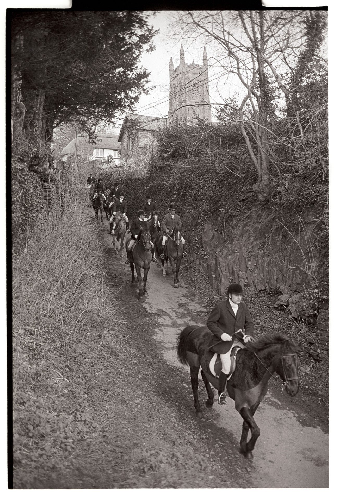 Hunt meet in front of Church, setting off down steep hill. 
[A hunt riding off down Rock Hill in Chulmleigh, to start hunting, after their Meet outside Chulmleigh Church, which is visible in the background.]
