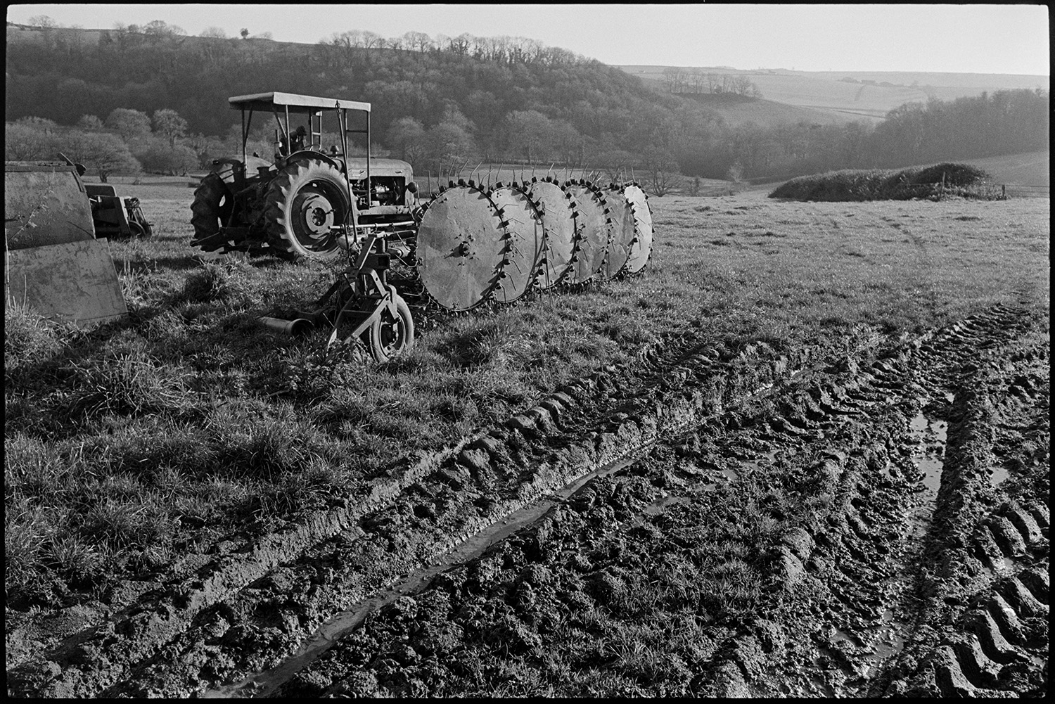 Tractor, muck spreader and whisk parked in muddy field.
[A tractor and hay whisk in a muddy field near Brushford. A wooded valley is visible in the background.]