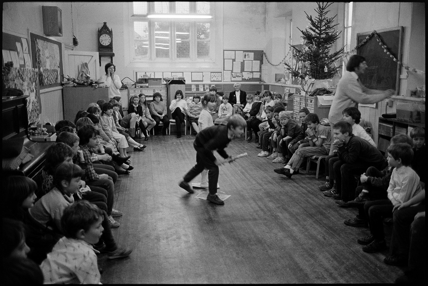 Christmas end of term party in primary school, games, teacher talking to pupils.
[A Christmas end of term party at Chulmleigh Primary School. The teachers, including Mr Sampson and Mrs Harris are organising games for pupils in the classroom, who are sitting on chairs around the room. There are paper chains, a Christmas tree, and decorations around the classroom.]