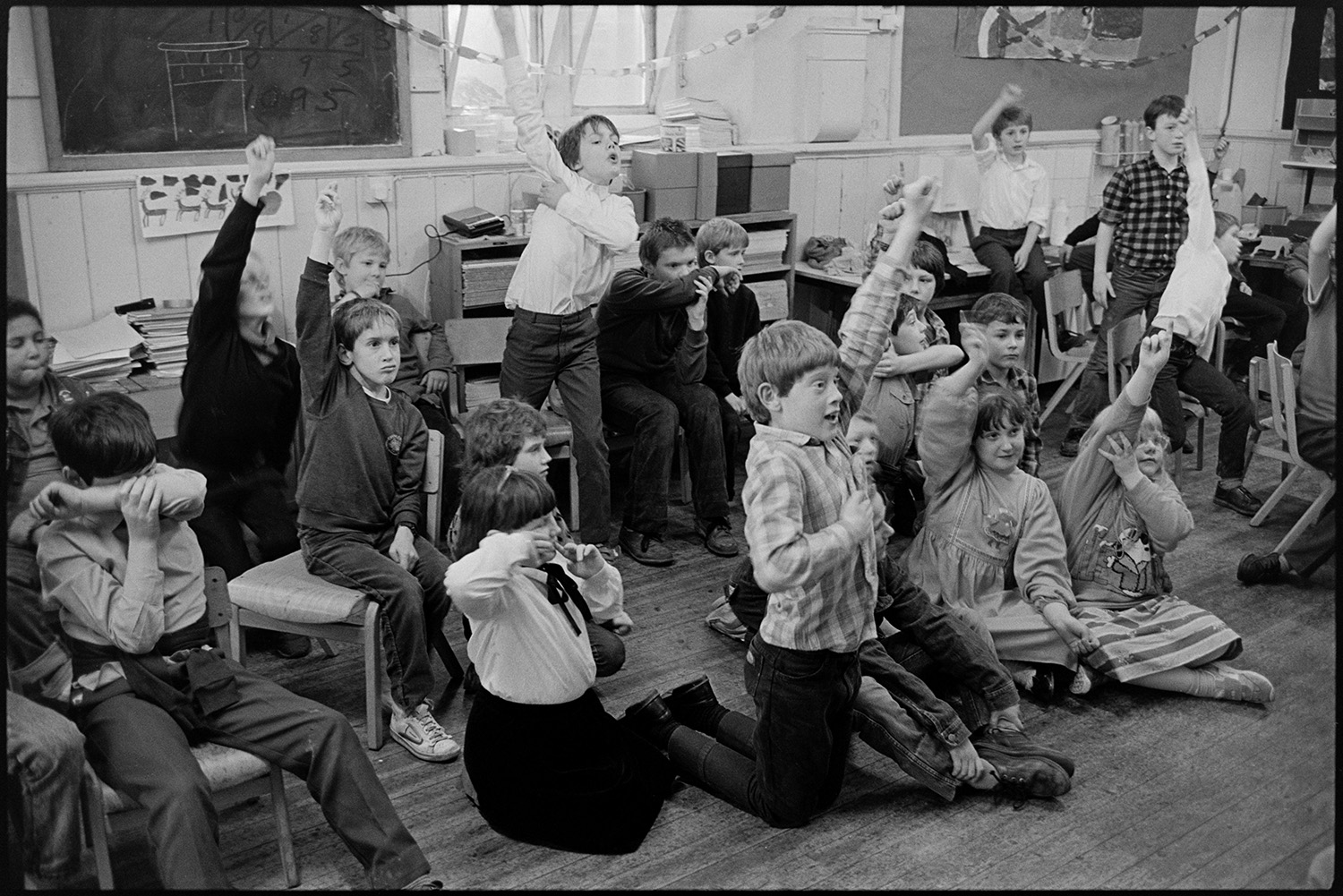 Christmas end of term party in primary school, games, teacher talking to pupils.
[A Christmas end of term party at Chulmleigh Primary School. The children who are sitting on chairs or on the floor, most with their hands up, are playing a party game. There are paper chains alongside blackboards, noticeboards and shelves of books in the classroom.]