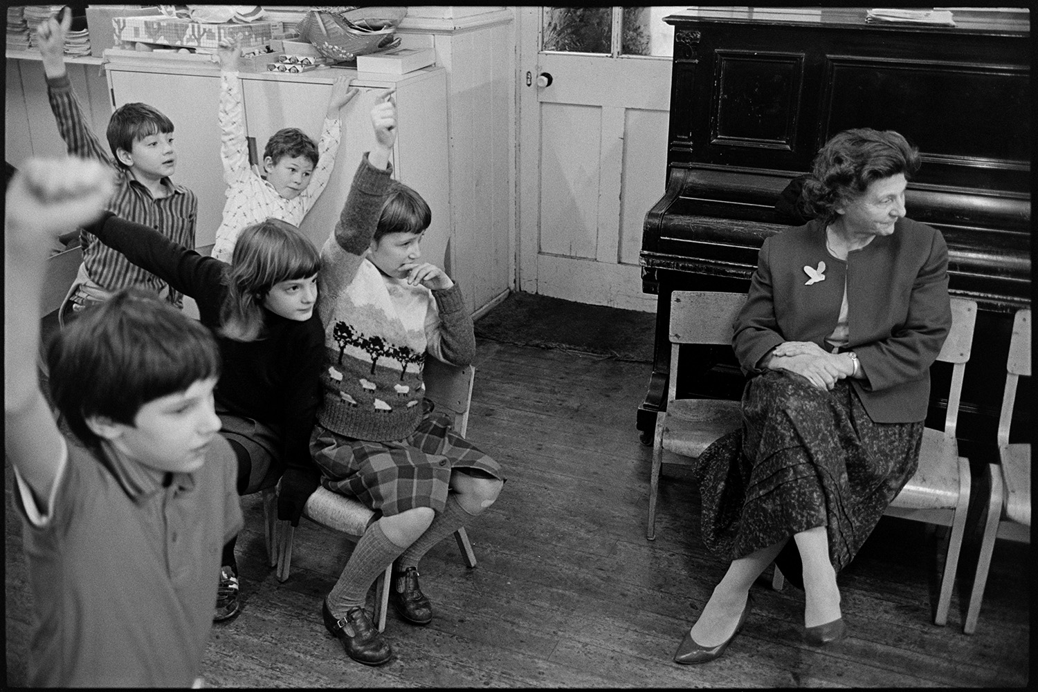 Christmas end of term party in primary school, games, teacher talking to pupils.
[A Christmas end of term party at Chulmleigh Primary School. The teachers are organising games for the pupils in the classroom, who are sitting on chairs with their hands in the air. A woman is sitting on a school chair in front of a piano.]