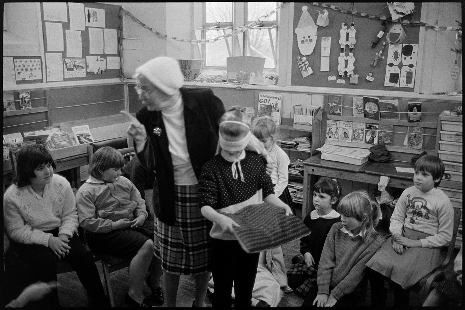 Christmas end of term party in primary school, games, teacher talking to pupils.
[A Christmas end of term party in a classroom at Chulmleigh Primary School. The children are playing party games and a woman is guiding a young pupil who is blindfolded and carrying a cushion, while other pupils sit and watch. There are paper chains and decorations around the classroom, alongside noticeboards, desks and shelves of books.]