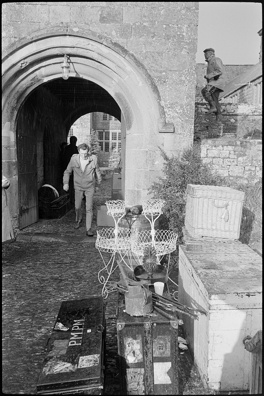 Large house sale, people looking at sale goods, watching bidding, panelled room, arch.
[Items put outside on a cobbled pathway for an auction at Colleton Manor, Chulmleigh, including metal chests, metal planters and a wicker chest. A man is walking through a stone archway, and another man is walking up some stone steps, in the background.]