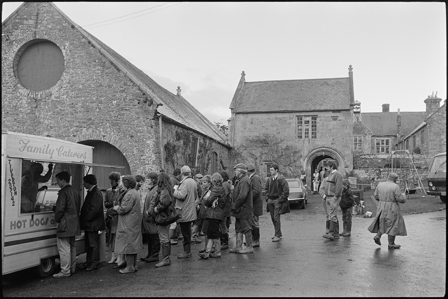 Large house sale, people looking at sale goods, watching bidding, panelled room, arch.
[Men and women lining up for refreshments at a catering van during an auction at Colleton Manor, Chulmleigh. Items for sale and stone buildings are visible in the background.]