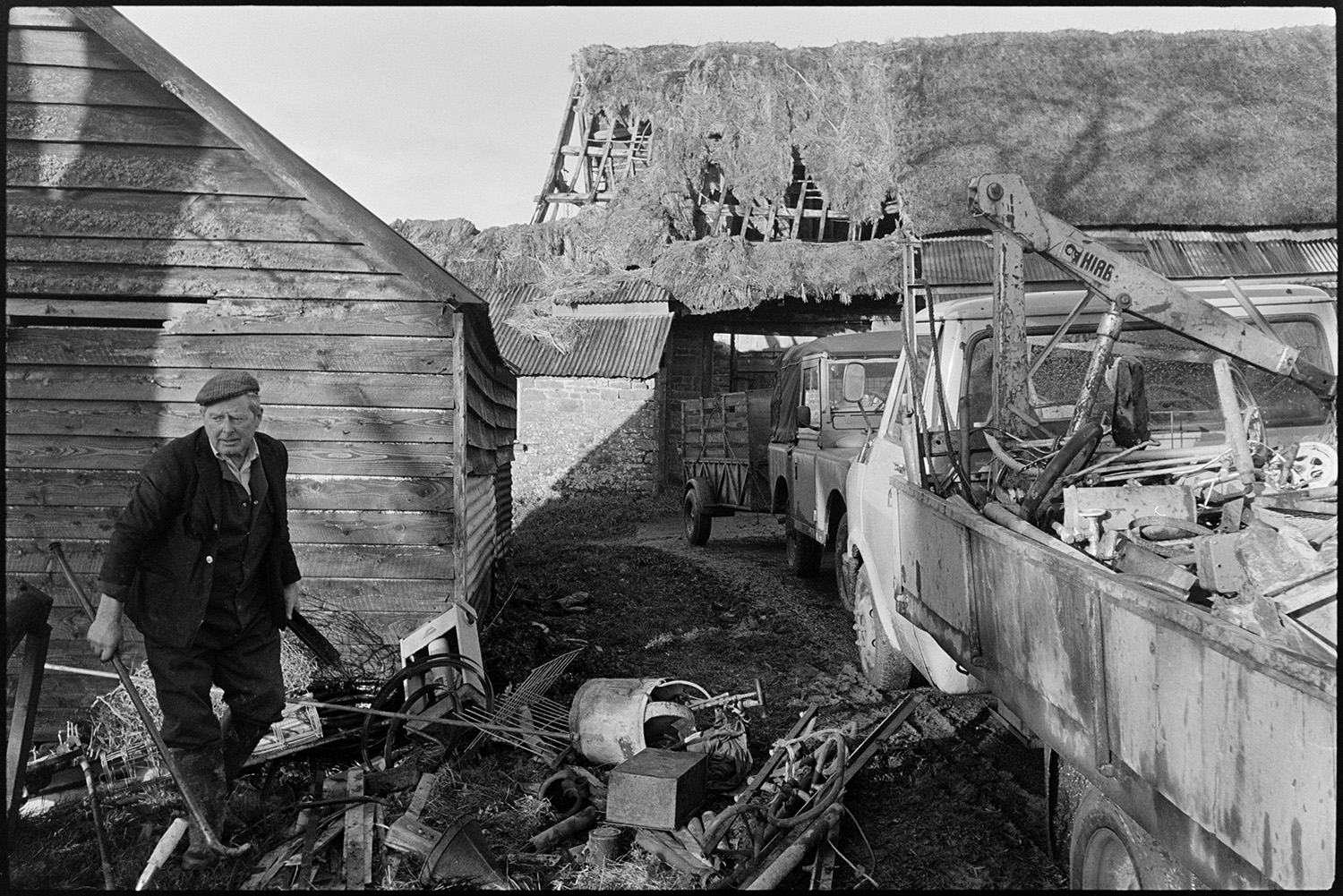 Scrap merchant sorting out scrap at country house sale.
[Scrap merchant Jimmy Hughes sorting out scrap materials at an auction at Colleton Manor, Chulmleigh. He is stood next to a wooden shed and a parked van, Land Rover and trailer. Stone barns with corrugated iron roofing and a collapsing thatched barn are visible in the background.]