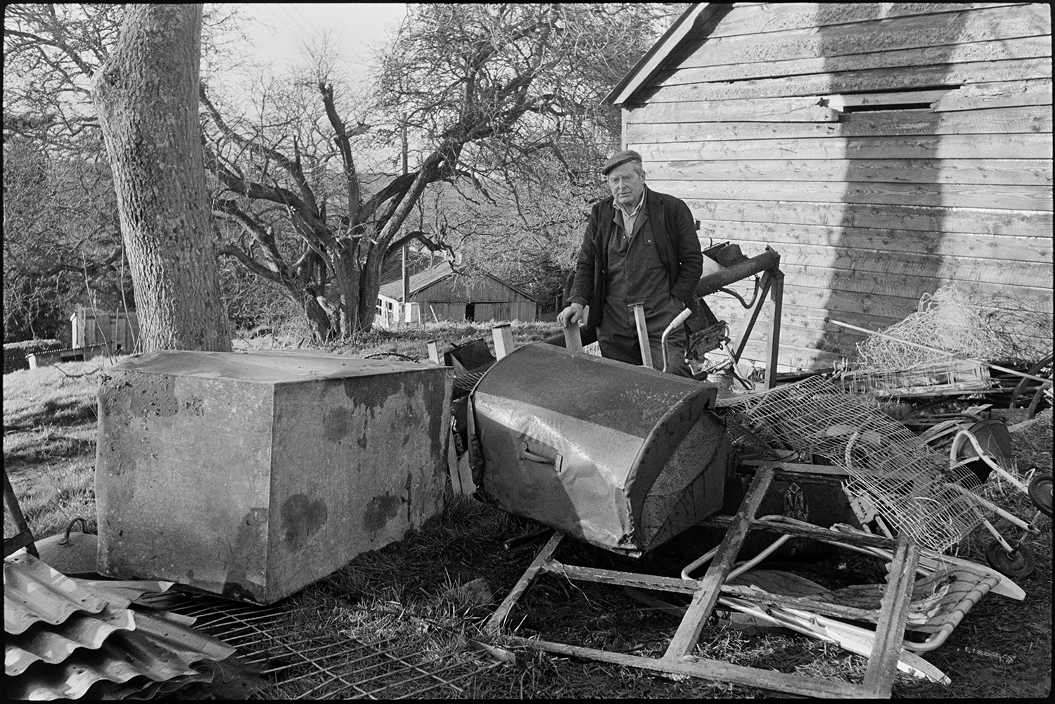 Scrap merchant sorting out scrap at country house sale.
[Jimmy Hughes, a scrap merchant, looking at a pile of scrap metal items including a bin, a tank and a window frame, at a sale at Colleton Manor, Chulmleigh. In the background is a large wooden barn.]