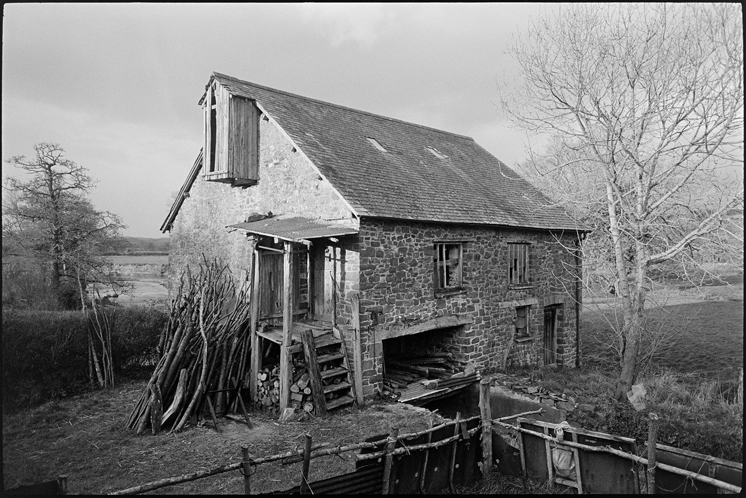 Mill building with woodpile,
[Rashleigh Mill, Ashreigney with a woodpile leaning against the wall. The stone mill building is surrounded by trees and fields.]