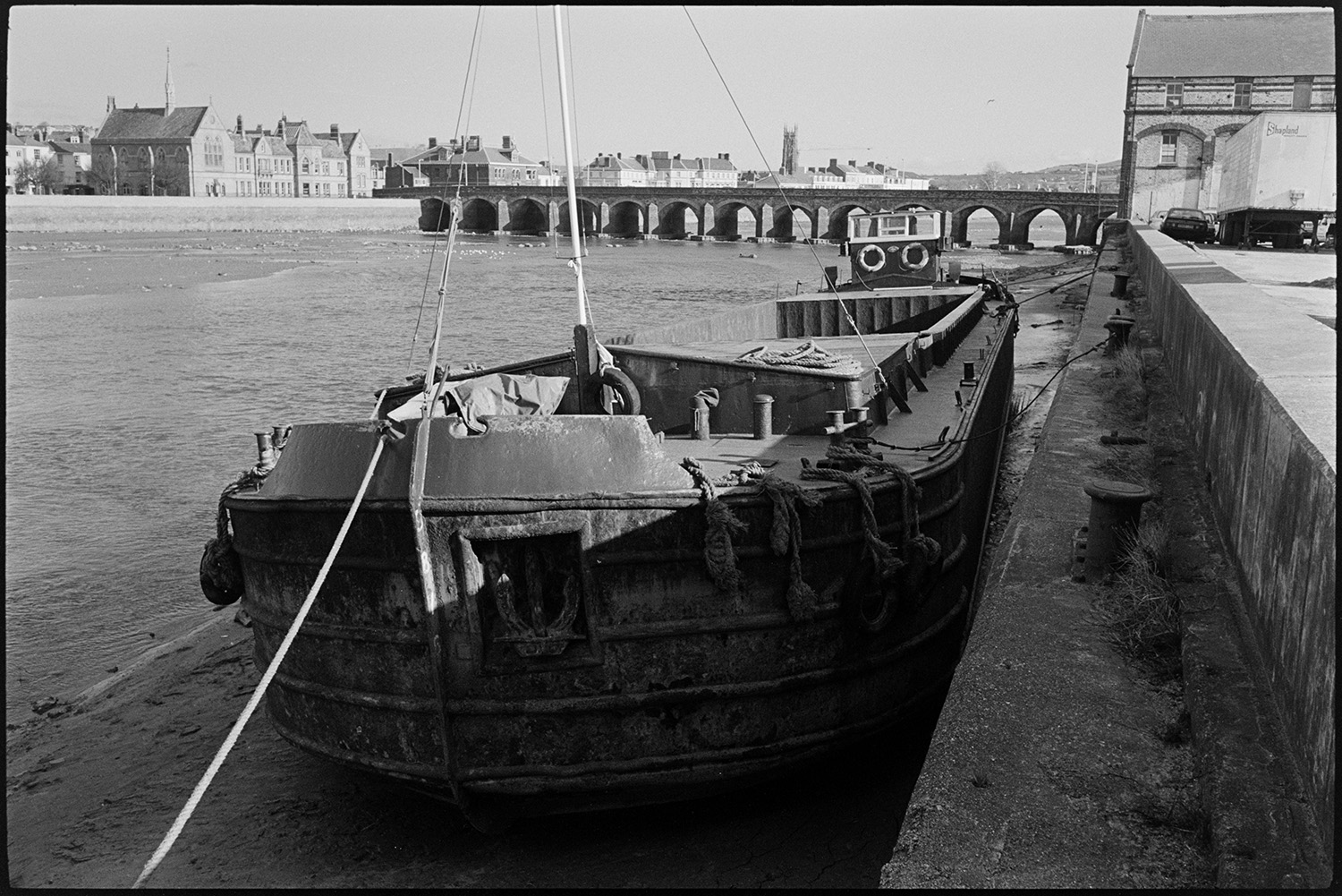 Barges and small boats on river bridge in background.
[A barge moored along the banks of the River Taw at Barnstaple. Barnstaple bridge and buildings along the river front can be seen in the background.]