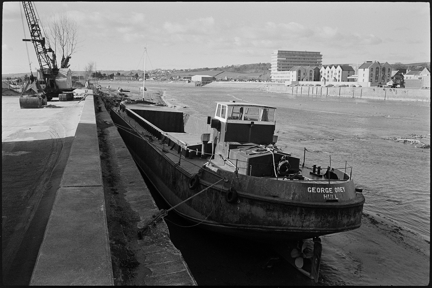 Barges and small boats on river bridge in background.
[Barge with the name George Odey, Hull moored to the bank of the River Taw, just to the west of Barnstaple. A crane with a loading grab is visible on the wharf. Large buildings and houses can be seen on the opposite side of the river.]