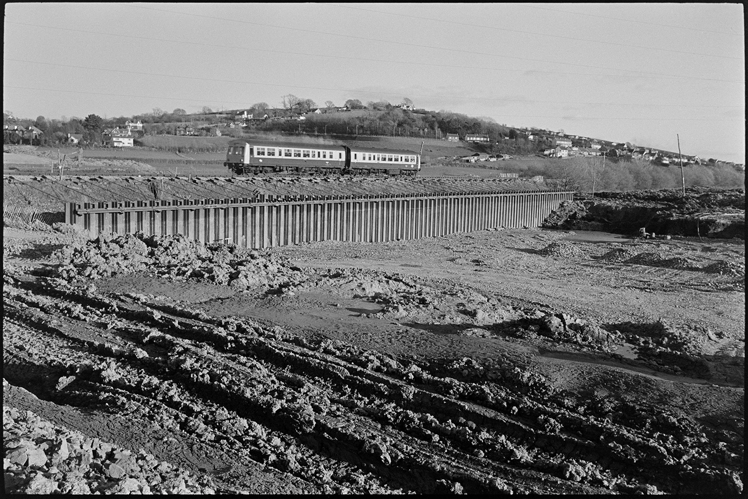Engineers working on new river bridge for motorway link road.
[A train on the Barnstaple to Bideford railway line passing between the River Taw and the preparatory work involved in the construction of a bridge over the river, near Barnstaple,]