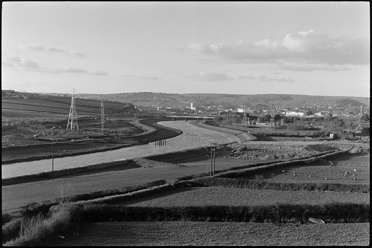 Engineers working on new river bridge for motorway link road.
[Piles in the River Taw being used in the erection of a bridge across the river. The town of Barnstaple can be seen in the background and electricity pylons are visible in the fields in the foreground.]