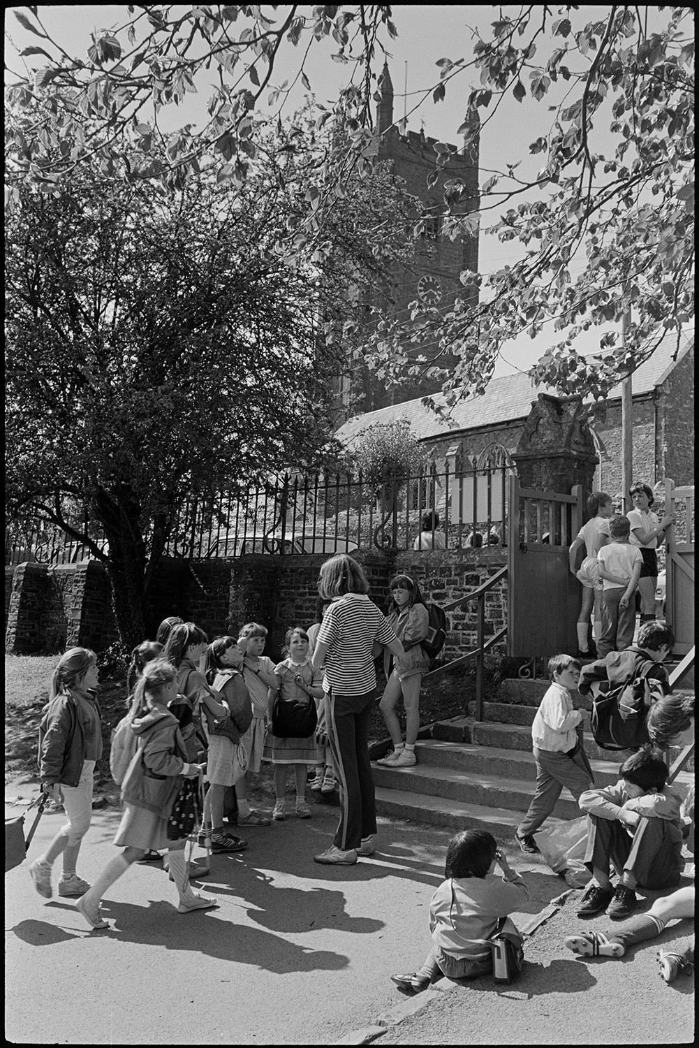 Children coming out of school. Going through cobbled streets in crocodile. Church tower.
[Children waiting and chatting at the steps by the school gate of Chulmleigh Primary School, opposite the Church which is visible in the background. Rowena Hoare, a teacher, is talking to the children.]