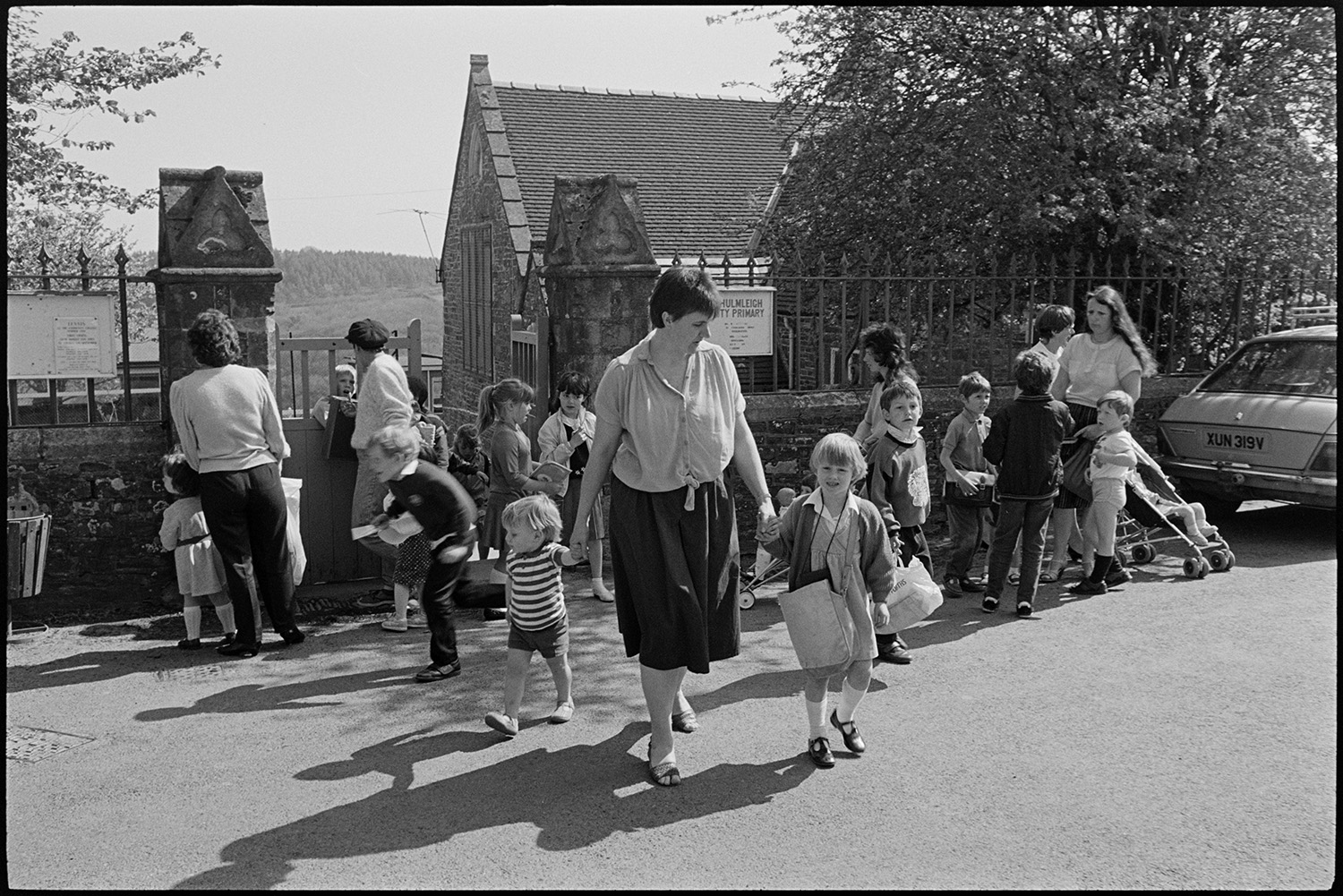 Children coming out of school. Going through cobbled streets in crocodile. Church tower.
[Mothers and women collecting children at the school gate, at Chulmleigh Primary School.]