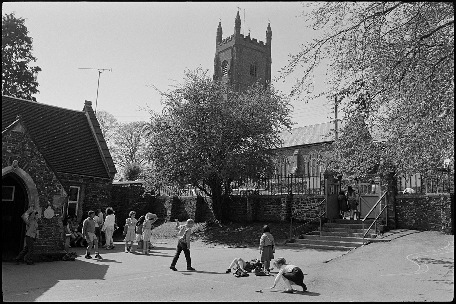 Children coming out of school. Going through cobbled streets in crocodile. Church tower.
[Children in the playground at the end of the school day at Chulmleigh Primary School. Some children are walking up the steps to meet their parents at the gate across the road from the church, which is visible in the background.]