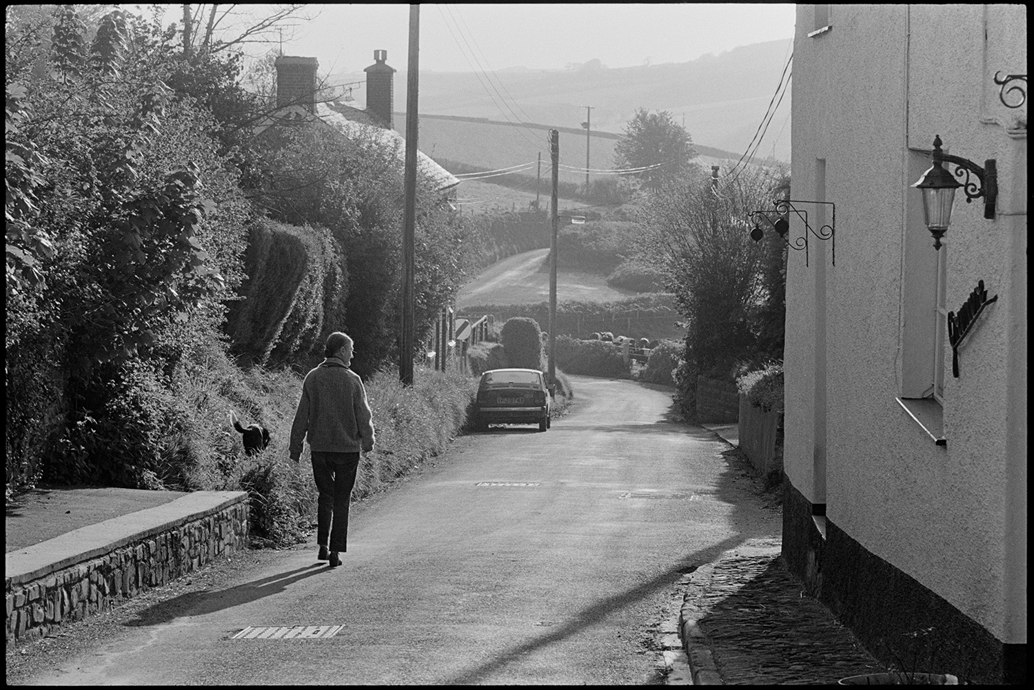Early morning, postman's round. Dustbin refuse bags in street. Cars passing and pony.
[A woman walking down the hill with her dog in morning sunshine at East Street, Chulmleigh. Se is passing a house with brackets for hanging baskets outside. A car is parked further along the street.]