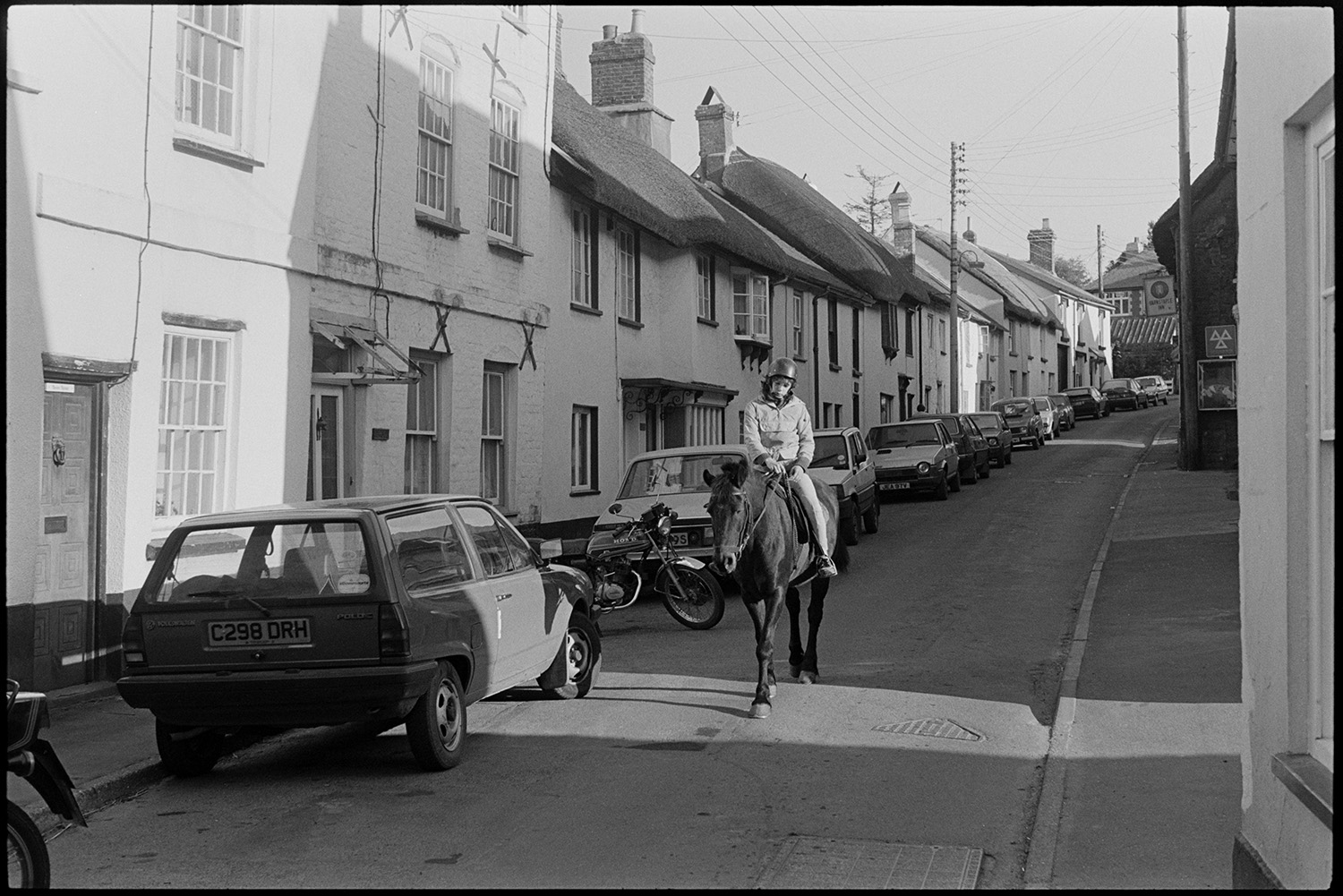 Early morning, postman's round. Dustbin refuse bags in street. Cars passing and pony.
[A young person a pony down East Street, Chulmleigh past the Barnstaple Inn, in the early morning. Parked cars and a motorbike are visible along the street, outside thatched cottages.]