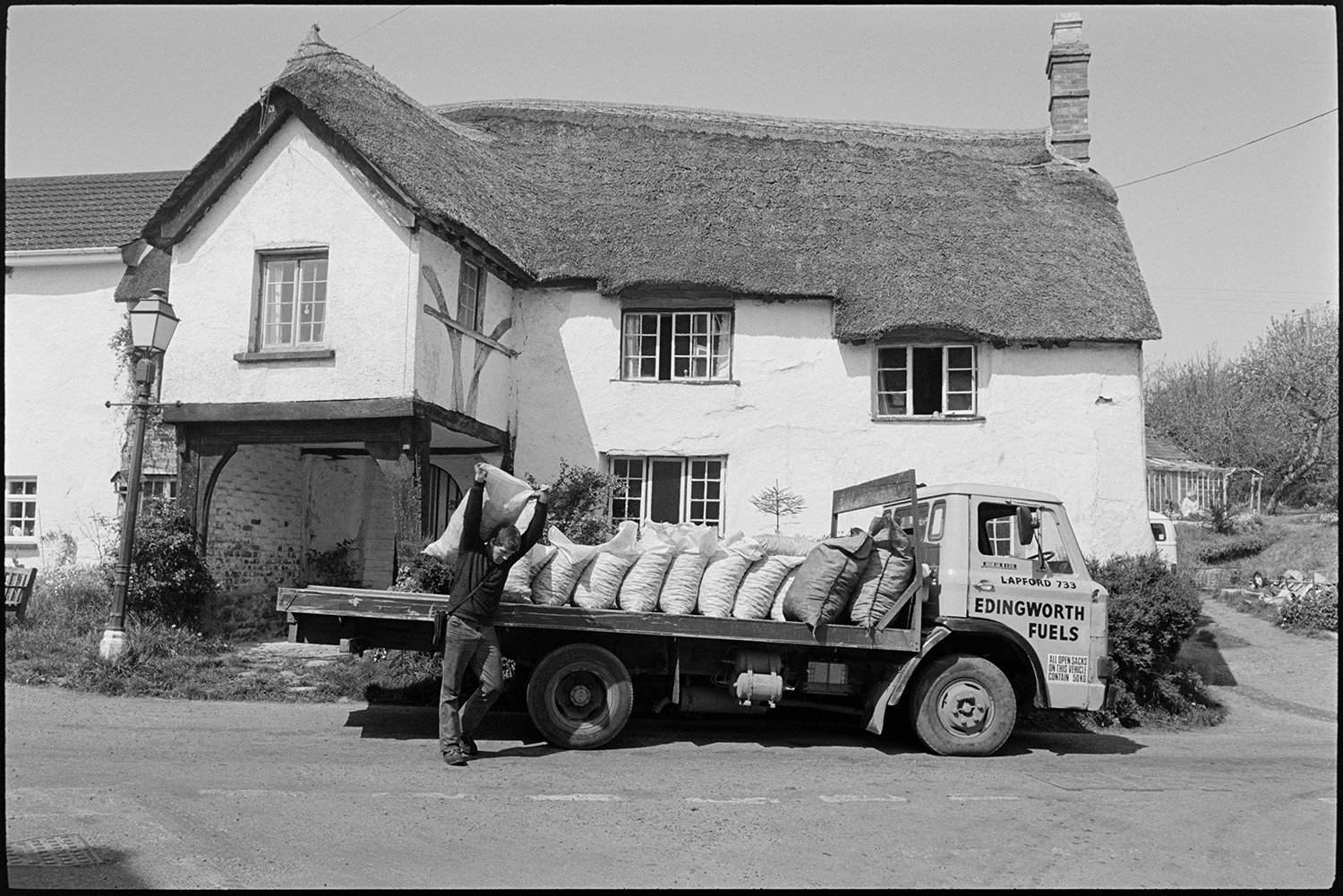 An Edingworth Fuels lorry parked by a thatched house with a lamp and a porch,  at Coleford.  A man is carrying and delivering sacks of coal from the lorry.