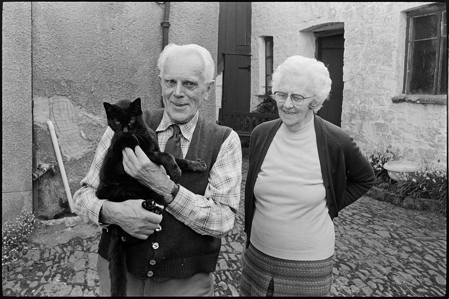 Man and woman. Wife standing with their pet cat.
[Ron Jury and Winnie Jury standing on the cobbled path outside their house at Leigh Road, Chulmleigh. Ron holding their pet cat.]