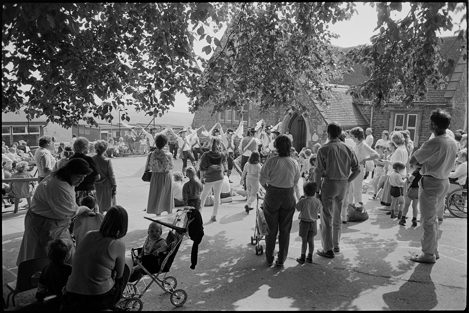 School fete, Morris dancers, stalls, former teacher chatting, bouncy castle, crowds.
[People gathered in the playground at Chulmleigh Primary School to watch Morris dancers at the school fete. Mothers and children are in the shade of a tree in the foreground.]