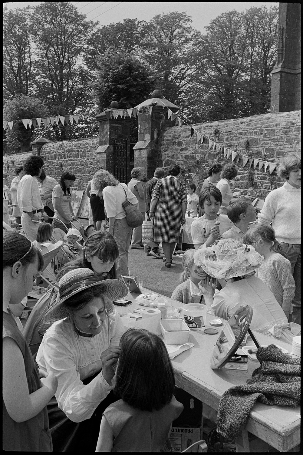 Chulmleigh Primary school fete with stalls, hand bells, eggs, games.
[People at Chulmleigh Primary School Fete looking at stalls.  Women wearing hats are painting children's faces on a stall in the foreground.]