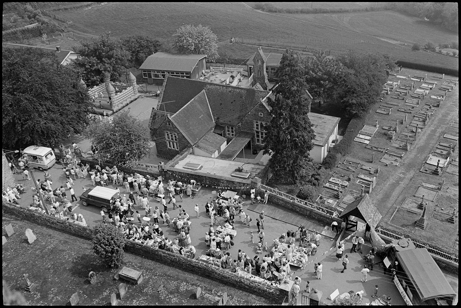 School fete viewed from Church tower. Helicopter flypast. Crowd. Former teacher chatting.
[A view from the church tower of Chulmleigh Primary School Fete and the graveyard. People visiting the fete in the playground are visible along with the school buildings.]