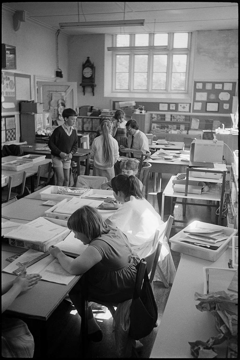 Primary school. Teacher helping pupils. Children working outside in hot sun.
[Richard Sampson, Headmaster, working with children in a classroom at Chulmleigh Primary School. The children's work is displayed around the room.]