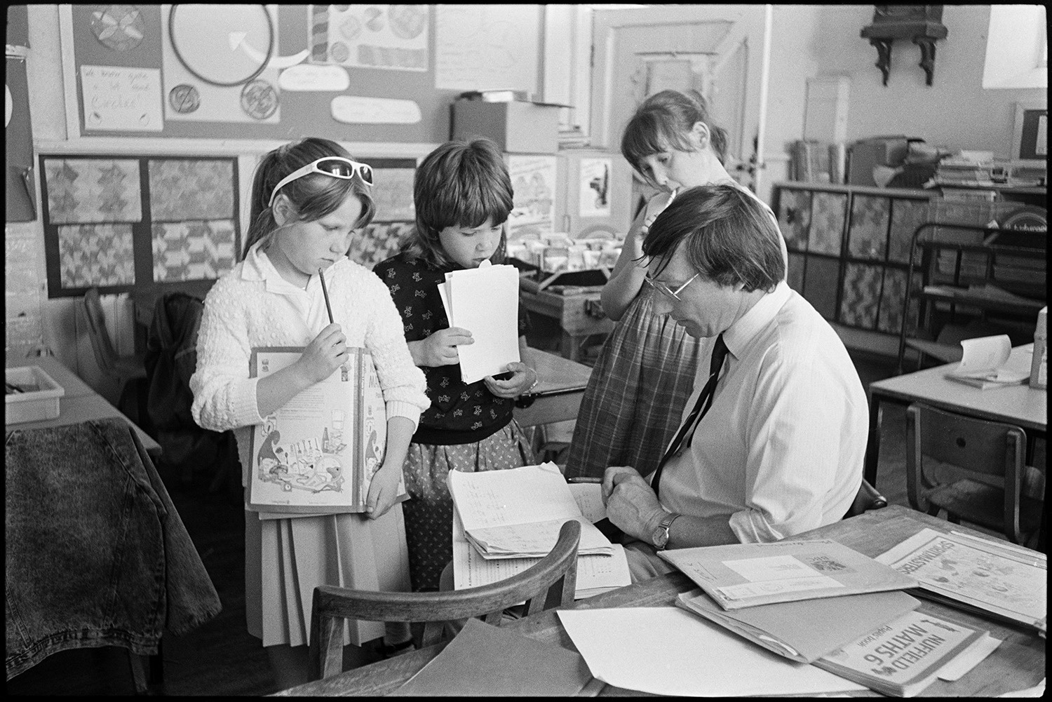Primary school. Teacher helping pupils. Children working outside in hot sun.
[Richard Sampson, Headmaster, working with children in the classroom at Chulmleigh Primary School. Desks and displays of children's work can be seen in the background.]