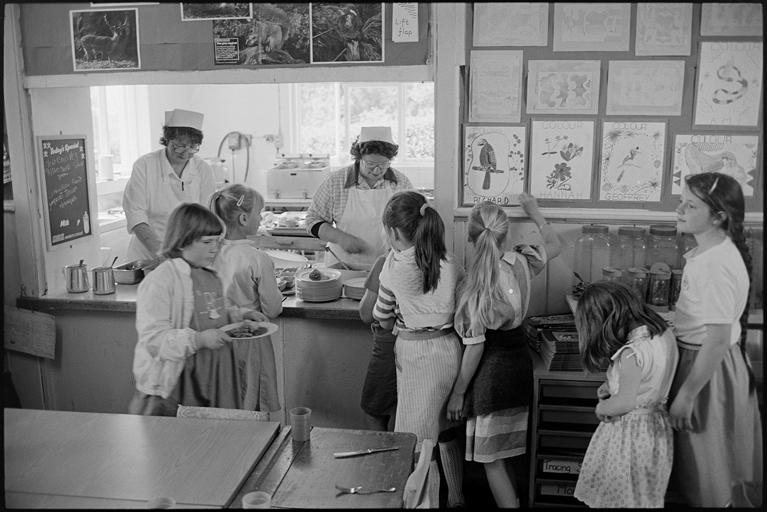 Primary school day. Teachers and pupils. Catering women making and serving lunch. Line up in yard.
[Two dinner ladies serving lunch to children from the canteen at Chulmleigh Primary School. A display of children's work is next to the serving hatch.]